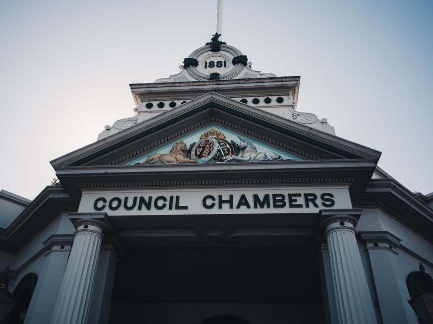 The image shows the façade of a historic council chambers building with classical architectural features, including two large white columns supporting a triangular pediment with decorative elements and a crest at the center. Above the pediment, there is a circular clock with a white face and black numerals, displaying the year 1881. The building's exterior is painted white with detailed molding and trim. The sky in the background is clear, with natural daylight illuminating the structure. The photograph captures a low-angle view, emphasizing the height and grandeur of the council chambers, which is relevant for a page about Camden Council permits for house removals and moving logistics, reflecting the administrative aspect of relocation planning as managed by local authorities like Man with van Frognal.