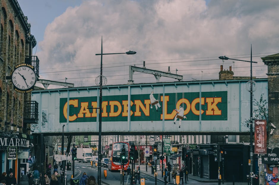 A wide view of Camden Lock in London, showing the iconic signboard with bold yellow and red lettering on a green background hanging over the street. Below the sign, pedestrians walk along the pavement while vehicles, including a red double-decker bus, travel through the busy area. The scene is set during daytime with partly cloudy skies overhead. To the left, part of an older brick building with a clock and balconies is visible, alongside shopfronts. In the middle distance, various street furniture, traffic lights, and street lamps are present, supporting the urban setting typical for home relocation and furniture transport planning. The image captures the vibrant atmosphere of a popular shopping and entertainment district, illustrating the importance of careful planning and logistics often handled by local removal and moving services, such as Man with van Frognal, especially when coordinated with Camden Council permits for street access and parking during house moving processes.