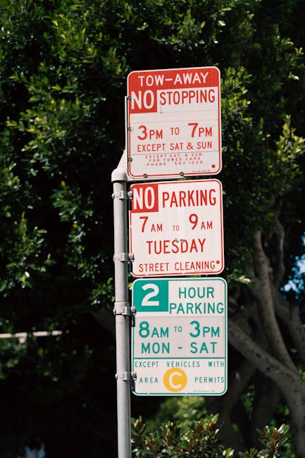 A set of three traffic and parking regulation signs mounted on a metal pole outdoors. The top sign is red and white, indicating a tow-away zone with no stopping permitted from 3pm to 7pm, except on Saturdays and Sundays, with additional contact details. Below it, a second sign in red and white specifies no parking from 7am to 9am on Tuesdays for street cleaning. The third sign is green and white, providing a two-hour parking limit from 8am to 3pm Monday to Saturday, with an exception for vehicles with area permits, designated by a yellow C symbol. The signs are situated in front of a leafy background with trees and blue sky, and the pole is positioned on a paved sidewalk, typical for urban street parking and traffic management in Frognal. This image is relevant to local rules and regulations concerning house removals, parking, and transport logistics in the area, as referenced on the page titled 'Do I Need a Skip or Permit in Frognal?' by Man with van Frognal.