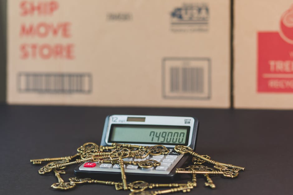 A small desk calculator with a green digital display showing the number 749800 is surrounded by multiple golden keys of various sizes, scattered across the black surface of the table. In the background, there are prominent cardboard boxes with red and blue text, indicating items for moving or storage, positioned inside a room or warehouse environment. The scene depicts a setting related to packing or moving logistics, with the objects arranged in front of large, labeled boxes, suggesting preparation for a house relocation or furniture transport service. The image emphasizes the aspects of packing materials and the logistical elements involved in home removals, aligning with cleaning and moving services performed by Man with van Frognal.