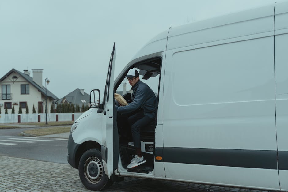 A man seated in the driver's seat of a white delivery van, with the driver's side door open, is holding a cardboard box in his hands during a home relocation process. The van is parked on a residentia