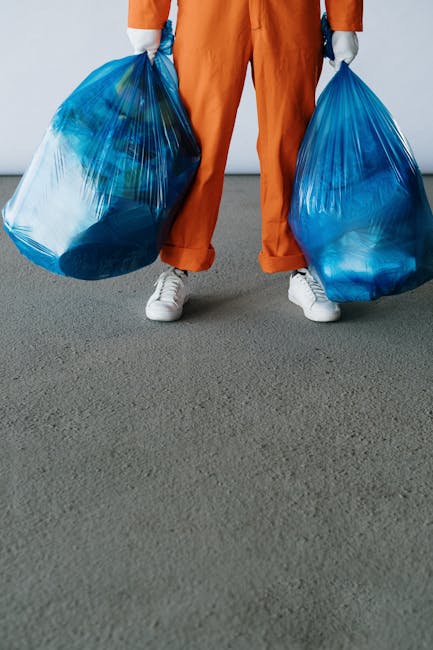 The image shows a person standing on a grey carpeted indoor floor, holding two large blue plastic trash bags filled with household waste or items ready for disposal, with the person's lower body visible. The individual is dressed in orange work trousers and white sneakers, and is wearing light grey gloves. Behind the person, part of a white wall and an open doorway are visible, suggesting an interior space likely being prepared for moving or clearance. This scene is associated with home relocation or packing and moving processes, illustrating the collection of waste or packing materials as part of a furniture transport or home removal activity. The overall environment appears clean and well-lit, emphasizing the task of loading or disposal during a house removal, possibly managed by a moving service such as Man with van Frognal, which specializes in removals and related logistics.