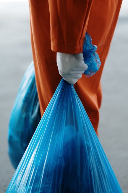 A person wearing an orange protective suit and white gloves is holding a blue plastic trash bag filled with waste, standing on a paved outdoor surface. The background is blurred, suggesting an outdoor environment, possibly near a building or loading area. The individual appears to be involved in waste disposal or cleanup activities, which may be part of a property clearance process related to home removal services. The image emphasizes careful handling of waste materials within a moving or relocation context, with the focus on the person's hand gripping the garbage bag and the surrounding setting indicating a step in packing or waste disposal before a house move.