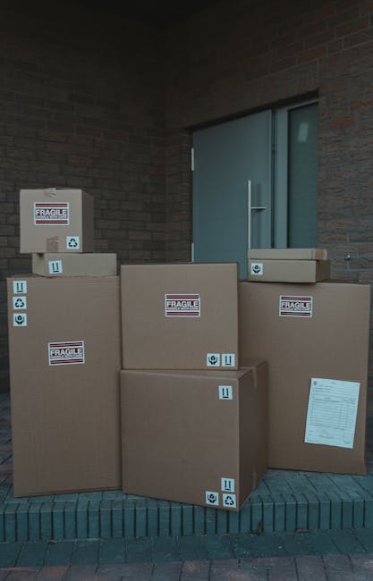 A man loading several cardboard boxes onto a white moving van parked on a residential street during daylight. The boxes are stacked on the pavement near the open rear door of the van, which is partial