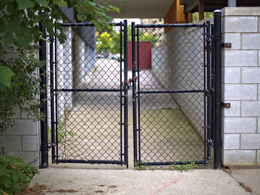 A black metal chain-link gate with two panels secured by a padlock, positioned between concrete block walls. The gate opens into a narrow passageway with a weathered concrete surface leading to a residential driveway. To the left, green foliage partially obscures the wall, while on the right, the concrete wall extends upward with visible mortar joints. The area appears clean and well-maintained, suitable for loading and unloading furniture during a home relocation. The surrounding environment indicates an exterior setting, with natural daylight illuminating the scene, supporting the process of packing and moving by [COMPANY_NAME], such as loading items into a van or preparing for furniture transport in a house removal.