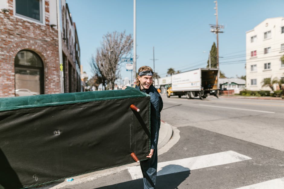 A man wearing a dark jacket and a patterned headband is standing outside on a street near a corner, holding a large piece of furniture wrapped in black fabric with orange handles. The furniture appears to be part of a house removal, as it is likely a mattress or similar item. Behind him, a white building with arched windows and a brick structure is visible on the left, while on the right, there are white multi-storey residential buildings and a utility pole with overhead wires. The street features a moving truck with an open back parked at the curb, and some cardboard packing materials may be present on the pavement. The scene is during daylight with clear skies, and the overall environment suggests a busy residential area prepared for home relocation, with [COMPANY_NAME] conducting furniture transport and packing activities as part of a professional removals service. The image captures the loading process, with the man smiling and ready to assist in the furniture transfer onto the truck or van, supporting efficient packing and house removals from Frognal Lane.