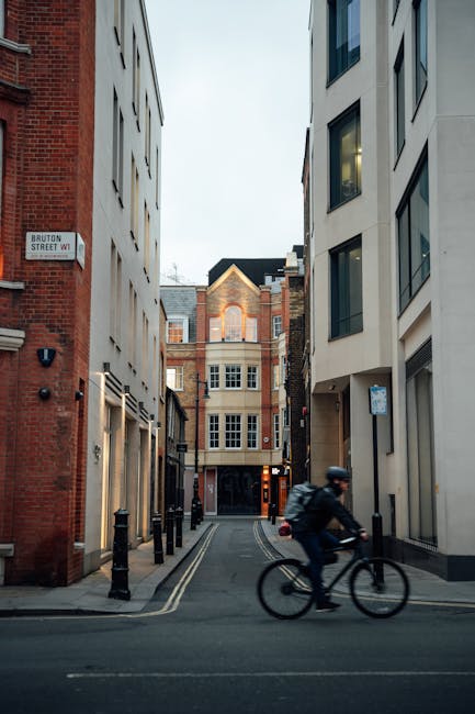 A cyclist wearing a dark helmet, jacket, and backpack is riding a bicycle along a narrow urban street with tall, modern and older brick buildings lining both sides. The street appears to be part of a dense city area, with the cyclist passing through a tight corridor of pavement and road markings. In the background, a building with white-framed windows and a peaked roof is visible, illuminated softly by natural daylight. Black bollards are positioned along the sidewalk edges, and a small street sign reading 'Bruton Street W1' is attached to a red brick building on the left. This scene depicts a typical city street environment, which may be relevant for house removals or local moving logistics, with a focus on the urban context and transportation passage. The image is well-suited for illustrating local relocations, furniture transport, or packing and moving processes often handled by companies like Man with van Frognal.
