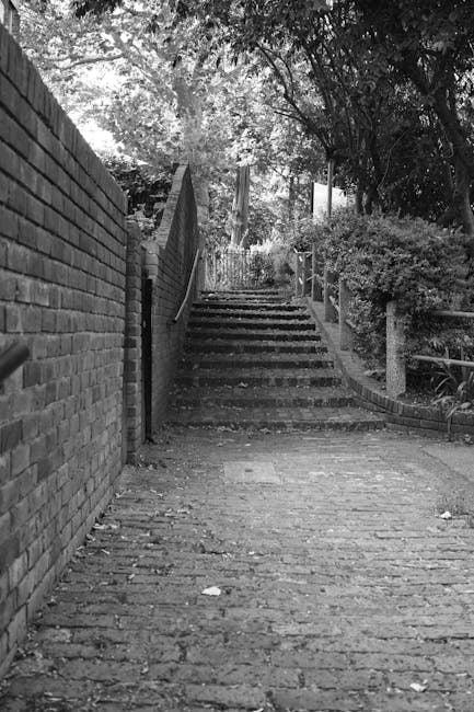 Black and white photograph of a narrow, outdoor stairway leading upward, composed of brick steps with visible wear. The stairs are flanked on the left by a high brick wall and on the right by a sloped, landscaped garden with bushes and trees. A metal railing runs along the right side of the stairs, which are partially shaded by overhanging tree branches. The area appears to be part of a residential or private property, with natural light filtering through the foliage. This setting illustrates a typical outdoor access route that may be encountered during a home relocation or furniture transport process, as managed by Man with van Frognal. The scene highlights the importance of planning for stair access during removals, especially in properties with limited outdoor space or steps to navigate before entering the home.