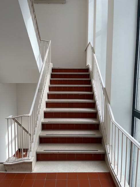Image of a well-lit indoor staircase inside a residential building, with white-painted metal handrails on both sides and polished wooden steps. The staircase has nine visible steps leading upward to an upper floor, with a white wall on the left and a large window on the right allowing natural light to illuminate the area. The floor at the base of the stairs is tiled with reddish-brown tiles, matching the stair treads. This interior space displays a clean and modern aesthetic, suitable for home relocation activities. Man with van Frognal offers professional removals services, including packing, furniture transport, and loading processes, often involving navigating staircases like this during house moves.