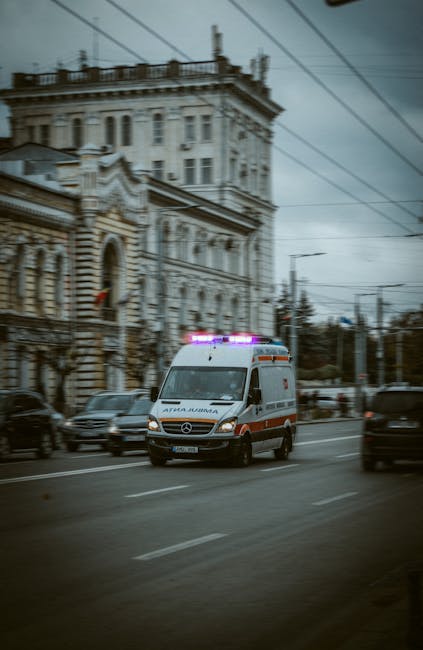 A white Mercedes-Benz ambulance with blue and red emergency lights flashing is driving along a city street under an overcast sky. The vehicle is positioned in the foreground, moving towards the viewer